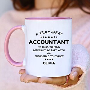 Può includere: Tazza in ceramica bianca con manico e interno rosa. La tazza presenta la scritta "A Truly Great Accountant" con il nome "Olivia" in basso. La tazza è tenuta da una persona.