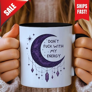 May include: White ceramic mug with a black interior and handle. The mug features a purple crescent moon design with stars and hanging crystals, and the text "DON'T FUCK WITH MY ENERGY".