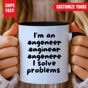 Puede incluir: Una taza de cerámica blanca con interior y asa negros, que presenta el texto en negro: I'm an engeneer, enginear, engenere, I solve problems, con las tres primeras palabras tachadas por líneas horizontales negras.