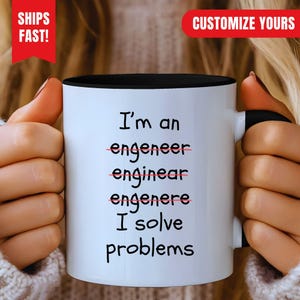 Może przedstawiać: Biały ceramiczny kubek z czarnym wnętrzem i uchwytem, z czarnym napisem: I'm an engeneer, enginear, engenere, I solve problems. Błędne pisownie są przekreślone cienką czerwoną poziomą linią.