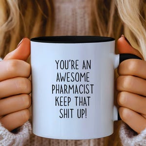 Op de afbeelding: Een witte keramische mok met een zwarte binnenkant en handvat. De mok heeft de tekst "YOU'RE AN AWESOME PHARMACIST KEEP THAT SHIT UP!" in het zwart, gecentreerd op de voorkant. De mok wordt door een persoon vastgehouden.