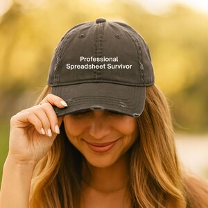 May include: A charcoal gray baseball cap with a distressed, worn-in finish. The front of the cap features the white embroidered text "Professional Spreadsheet Survivor" in a clean, sans-serif font. The cap has a curved brim and stitched panels.