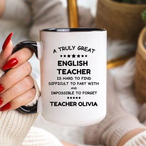 Puede incluir: Taza de cerámica blanca con asa y borde negros. La taza presenta el texto "A Truly Great English Teacher" con texto adicional debajo. La taza está personalizada con el nombre "Teacher Olivia".