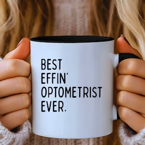 May include: A white ceramic mug with a black interior and handle. The mug features the text "BEST EFFIN' OPTOMETRIST EVER" in bold black letters. The mug is held by a person wearing a light-colored sweater.
