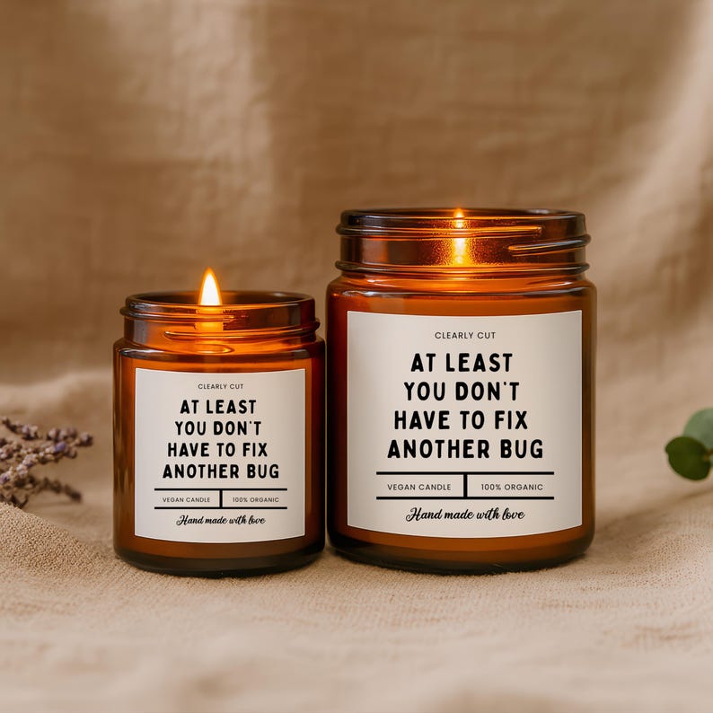 Peut inclure: Deux bougies en pot en verre ambr&eacute; avec des m&egrave;ches allum&eacute;es. Chaque &eacute;tiquette blanche porte le texte : CLEARLY CUT, AT LEAST YOU DON'T HAVE TO FIX ANOTHER BUG, VEGAN CANDLE, 100% ORGANIC, et Hand made with love. Les bougies sont de tailles diff&eacute;rentes.