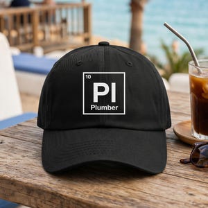 May include: A black baseball cap featuring a white square graphic on the front. Inside the square, the number 10 is in the top left corner, the letters PI are in the center, and the word Plumber is printed in white text at the bottom.