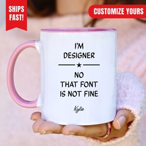 Könnte beinhalten: Ein weißer Keramikbecher mit einem hellrosa Henkel und einem hellrosa Innenrand. Der Becher trägt den schwarzen Schriftzug: I'M DESIGNER NO THAT FONT IS NOT FINE, mit einem Sternsymbol in der Mitte und dem Namen Kylie in Schreibschrift unten.