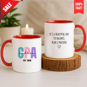 Könnte beinhalten: Zwei weiße Keramiktassen mit leuchtend roten Innenseiten und Henkeln. Eine Tasse zeigt den bunten Text CPA und andrea mit EST. 2026 darunter. Die andere Tasse zeigt den schwarzen Text IT'S A BEAUTIFUL DAY TO BALANCE, PLAN & PROSPER mit einem Herz.
