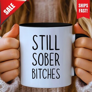 May include: White ceramic mug with a black interior and handle. The mug features the text "STILL SOBER BITCHES" in black, bold letters. A red banner in the top right corner reads "SHIPS FAST!". A red banner in the top left corner reads "SALE".
