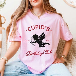 Puede incluir: Camiseta rosa claro con un diseño de Cupido y el texto ".CUPID'S. Birthday Club ESTD 1974". El diseño es en rojo oscuro y negro. La camiseta tiene cuello redondo y mangas cortas.