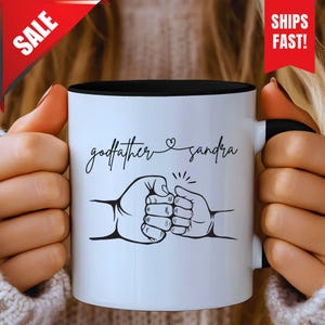 May include: White ceramic mug with a black interior and handle. The mug features the text "godfather & sandra" in cursive, along with a graphic of two fists bumping. A red "SALE" banner and a "SHIPS FAST!" banner are visible.