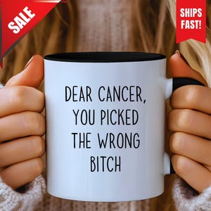 Puede incluir: Taza de cerámica blanca con interior y asa negros. La taza presenta el texto "DEAR CANCER, YOU PICKED THE WRONG BITCH" en letras negras y en negrita. Una pancarta roja "SALE" está en la esquina superior izquierda, y una pancarta roja "SHIPS FAST!" está en la esquina superior derecha.