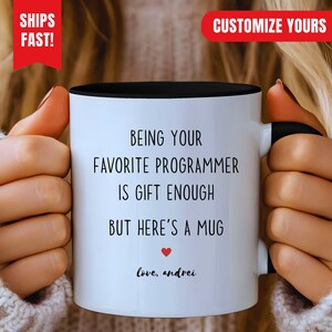 Könnte beinhalten: Ein weißer Keramikbecher mit schwarzem Innenraum und Henkel. Auf der Vorderseite steht in schwarzer Schrift: BEING YOUR FAVORITE PROGRAMMER IS GIFT ENOUGH BUT HERE'S A MUG, gefolgt von einem kleinen roten Herz und dem Text: love, andrei.