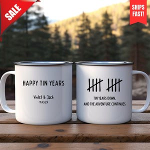 May include: Two white enamel mugs with black rims and handles. One mug reads "HAPPY TIN YEARS" with a name and date. The other mug has tally marks and the text "TIN YEARS DOWN, AND THE ADVENTURE CONTINUES."