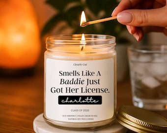 Peut inclure: Une bougie en pot de verre transparent avec une étiquette blanche portant le texte "Clearly Cut", "Smells Like A Baddie Just Got Her License.", "charlotte", "CLASS OF 2026", "ECO-FRIENDLY | VEGAN | MADE IN USA" et "NATURAL SOY WAX CANDLE".