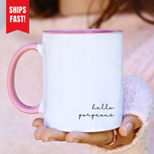 Può includere: Tazza in ceramica bianca con interno e manico rosa. La tazza presenta la scritta "hello gorgeous" in un carattere scritto a mano. La tazza è tenuta da una persona.