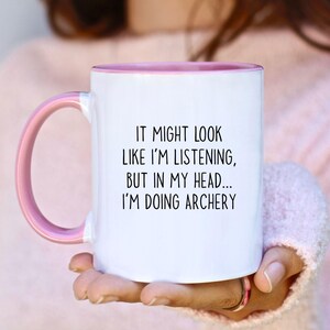 May include: White ceramic mug with a pink interior and handle. The mug features the text "IT MIGHT LOOK LIKE I'M LISTENING, BUT IN MY HEAD... I'M DOING ARCHERY" in black lettering. A fun gift for archers.