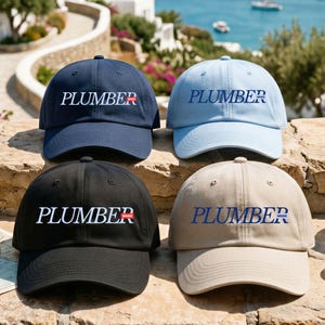 May include: Four baseball caps in navy blue, light blue, black, and beige. Each cap features the text PLUMBER MODE embroidered on the front. The navy and black caps have white text with a red box around the word MODE, while the light blue and beige caps have blue text.