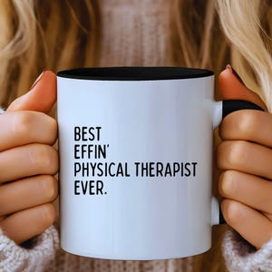 Puede incluir: Una taza de cerámica blanca con interior y asa negros. La taza presenta el texto "BEST EFFIN' PHYSICAL THERAPIST EVER" en letras negras y en negrita.