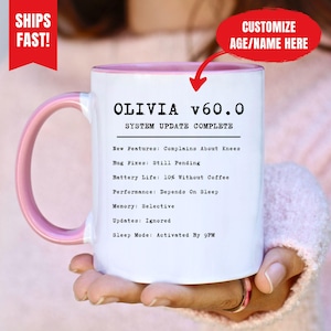 Op de afbeelding: Witte keramische mok met een roze binnenkant en oor. De mok heeft de tekst "OLIVIA v60.0 SYSTEM UPDATE COMPLETE" met humoristische updates zoals "Complains About Knees" en "Battery Life: 10% Without Coffee". Een rode banner leest "CUSTOMIZE AGE/NAME HERE".