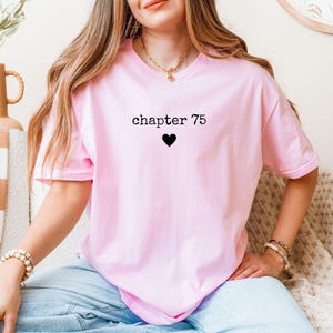 Puede incluir: Una camiseta rosa claro con el texto "chapter 75" encima de un corazón negro. La camiseta es de manga corta y parece estar hecha de un material suave. La persona lleva vaqueros azules y un collar dorado.