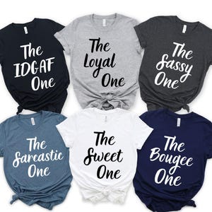 Puede incluir: Seis camisetas de manga corta en varios colores, cada una con un diseño de texto blanco diferente. Las camisetas incluyen las frases "The IDGAF One", "The Loyal One", "The Sassy One", "The Sarcastic One", "The Sweet One" y "The Bougee One."