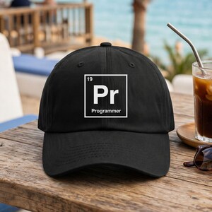 Puede incluir: Una gorra de béisbol negra con un gráfico cuadrado blanco en la parte delantera. Dentro del cuadrado aparecen el número 19, las letras Pr y la palabra Programmer, diseñados para parecer un elemento de la tabla periódica de elementos.