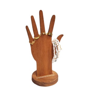 May include: A wooden hand-shaped jewelry display stand. The stand is brown and holds several gold rings and a beaded bracelet. The base of the stand is round, and the text "@THEARTOFDISPLAY" is printed on the base.