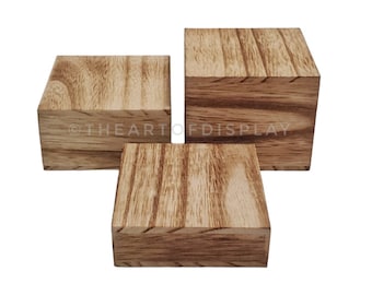 Puede incluir: Tres elevadores de exhibición de madera en varios tamaños. Los elevadores están hechos de madera marrón claro con un acabado de veta de madera natural. Los elevadores son de forma cuadrada y tienen una superficie lisa.