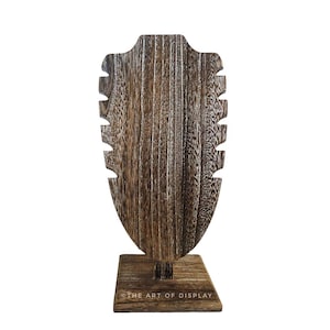 May include: Wooden necklace display stand with a brown wood finish and a rectangular base. The stand has a cutout in the shape of a neck to hold necklaces. The stand is labeled "The Art of Display".