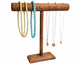 May include: A wooden jewelry display stand with a horizontal bar and a base. The stand holds several necklaces, including gold and blue beaded strands, and delicate pendant necklaces. The stand is made of dark wood and is set against a white background.