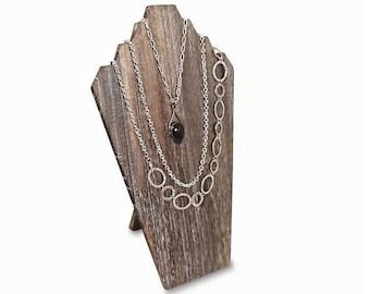 May include: A rustic wooden jewelry display stand with three silver necklaces. The necklaces have different designs, including a pendant and a chain of connected circles. The stand is made of weathered wood and has a natural, textured appearance.