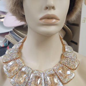 May include: A mannequin wearing a tan faux fur hat and a statement necklace. The necklace features large, rectangular, iridescent, amber-colored stones set in a sparkling, glittery frame. The necklace is a bold fashion accessory.