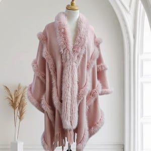May include: A pink knitted poncho with a faux fur trim. The poncho has a fringe detail and is displayed on a mannequin.
