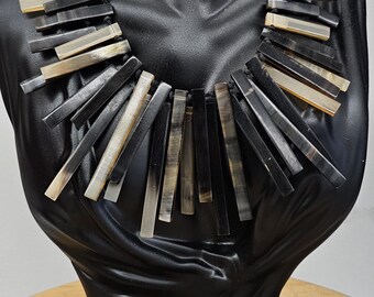 May include: A striking necklace composed of numerous rectangular, dark-colored pieces, interspersed with lighter, cream-colored segments. The necklace is displayed on a black bust, showcasing its design and texture. The jewelry is set against a neutral background.