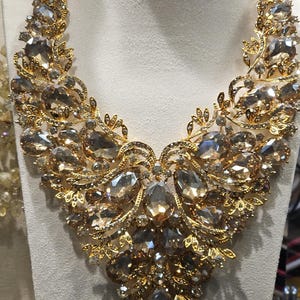 May include: A gold-toned statement necklace with large, faceted, amber-colored gemstones. The necklace features intricate filigree and leaf-like details, displayed on a white bust. A luxurious piece of jewelry.