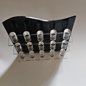 May include: A black acrylic display stand with two clear shelves and cylindrical holders. The top shelf is a curved shape. The stand is designed to hold multiple items, likely small bottles or vials.