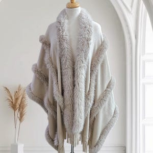 May include: A beige faux fur shawl with a long fringe. The shawl has a soft, fluffy texture and is trimmed with a thick band of faux fur.