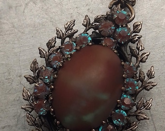 May include: A vintage brooch with a large, brown, oval-shaped cabochon stone surrounded by a ring of smaller, iridescent blue rhinestones. The brooch is set in a silver-toned metal with a leafy design.