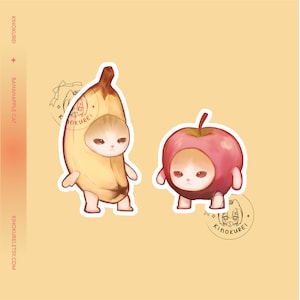 May include: Two adorable cartoon cats dressed as fruit. One cat is wearing a yellow banana costume and the other is wearing a red apple costume. Both cats have sad expressions on their faces.