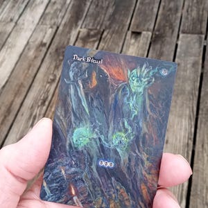 May include: A trading card with the text "Dark Ritual" at the top. The card features a dark, fantastical illustration with green and blue spectral figures and a fiery orange glow. The card is held in a person's hand.