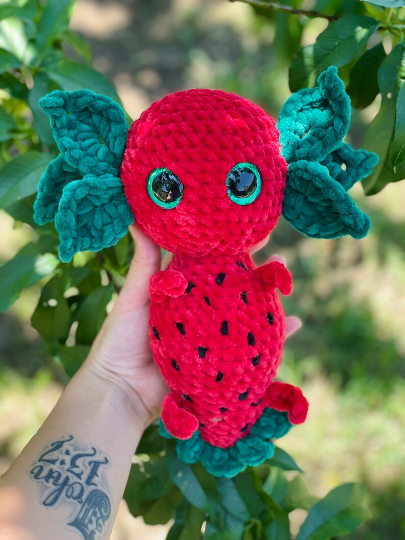 May include: A red and green crocheted axolotl plush toy with large black eyes. The axolotl is shaped like a strawberry with green leaves.