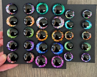 May include: Assortment of colorful, glittery googly eyes of various sizes, arranged on a black background. The eyes have a black pupil and a variety of colors, including green, blue, purple, orange, and pink.