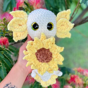 May include: A white crocheted axolotl plush toy with yellow crocheted gills and a sunflower. The axolotl has large black eyes and a brown crocheted nose.