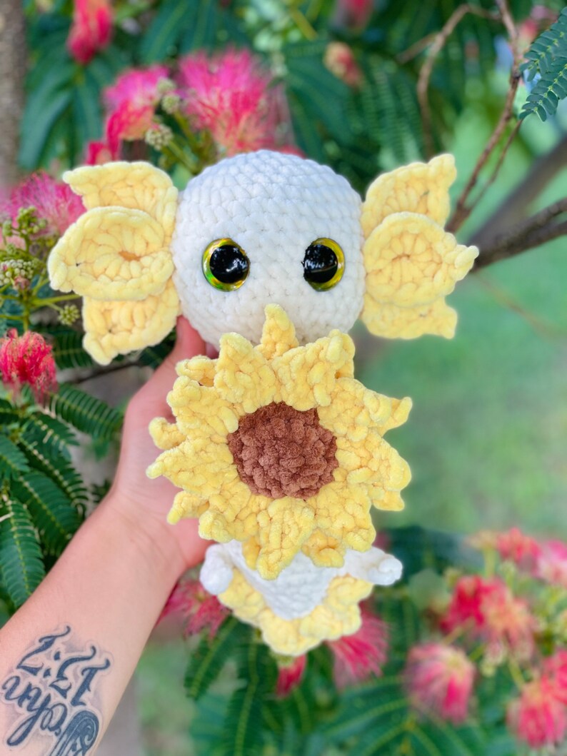 May include: A white crocheted axolotl plush toy with yellow crocheted gills and a sunflower. The axolotl has large black eyes and a brown crocheted nose.