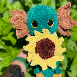 May include: A crocheted plush toy in the shape of a green axolotl holding a yellow sunflower. The axolotl has brown ears and black eyes with yellow pupils.