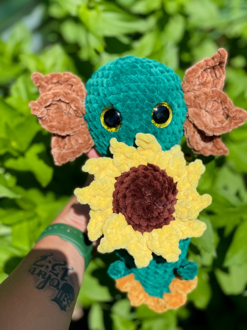 May include: A crocheted plush toy in the shape of a green axolotl holding a yellow sunflower. The axolotl has brown ears and black eyes with yellow pupils.