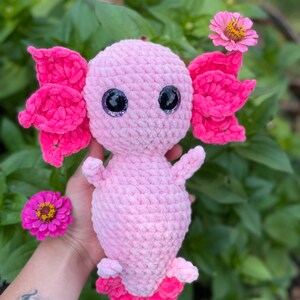 May include: Pink crocheted axolotl plush toy with large googly eyes. The toy is sitting in a hand with green foliage in the background.
