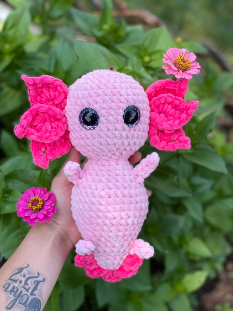 May include: Pink crocheted axolotl plush toy with large googly eyes. The toy is sitting in a hand with green foliage in the background.