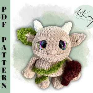 May include: A plush, beige crochet creature with large, colorful eyes and small horns. It has green accents on its ears and chest, and holds a burgundy heart. The image includes the text "PDF PATTERN" and a logo.